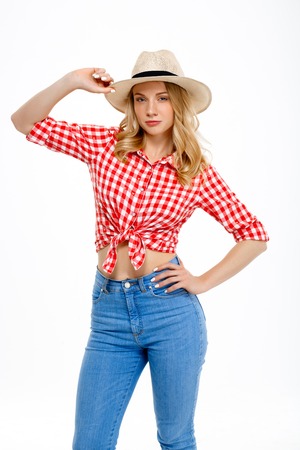 Portrait of young beautiful country girl in hat, jeans and shirt smiling, looking at camera over white background.の写真素材