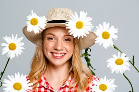 Portrait of young beautiful country girl in hat with chamomiles smiling, looking at camera over gray background.の写真素材