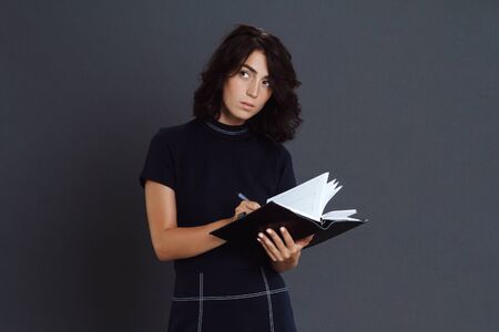 Thoughtful young woman posing over grey background and holding notebook in hands