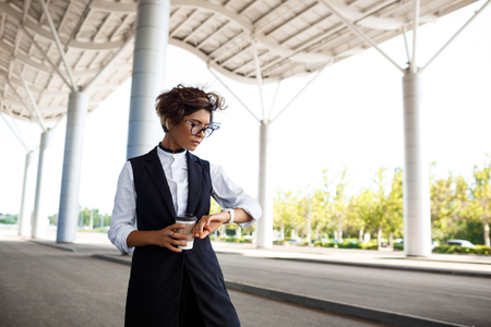 Young successful businesswoman in glasses looking at watch, holding coffee, standing near business centre.の写真素材