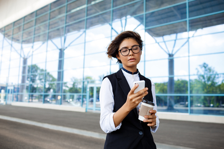 Young successful businesswoman in glasses looking at phone, holding coffee, standing near business centre.の写真素材