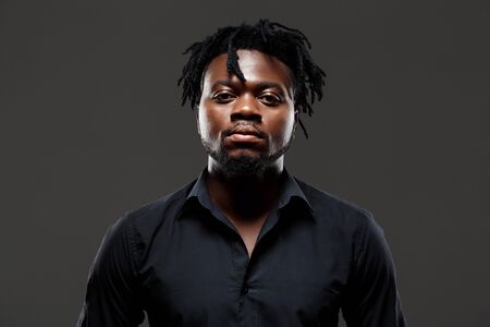 Young successful african businessman in black shirt posing, looking at camera over dark background....