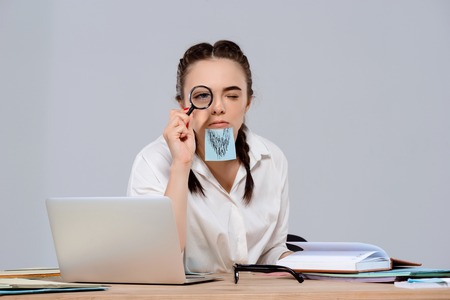 Young beautiful businesswoman looking at camera through magnifier, sticker on chin, sitting at workplace over purple background. Copy space.の写真素材