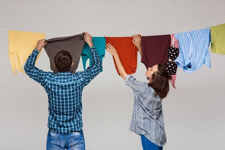 Young beautiful married couple hanging wash clothes over grey background. Copy space.