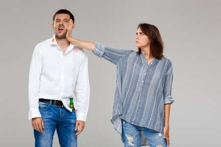 Young beautiful woman quarreling, angering with drunk husband over grey background. Copy space.