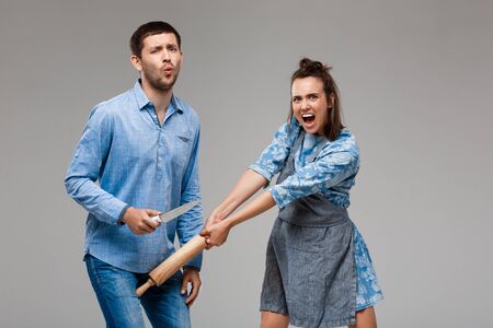Young beautiful woman beating man with rolling pin, shouting over grey background. Copy space.