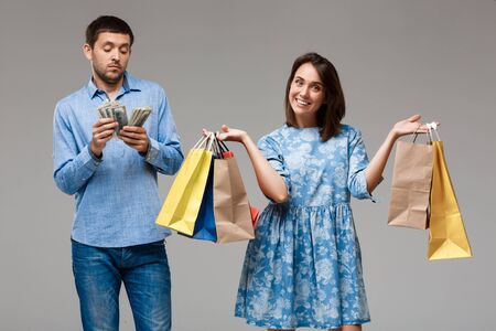 Young beautiful woman holding purchases, man holding money over grey background. Copy space.の写真素材