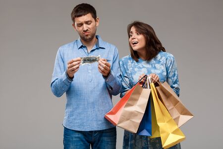 Young beautiful woman holding purchases, man holding last money over grey background. Copy space.の写真素材