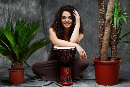 Young beautiful brunette girl smiling, sitting and playing drum in tropical plants over grey background.の写真素材