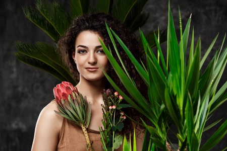 Young beautiful brunette girl in tropical plants looking at camera, smiling over grey background.の写真素材