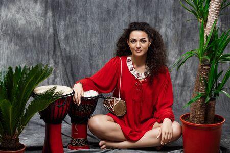 Young beautiful brunette girl smiling, sitting and playing drum in tropical plants over grey background.の写真素材