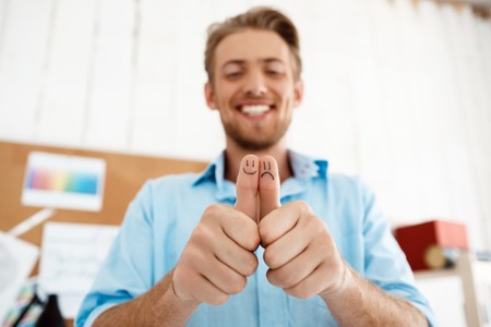 Young handsome smiling businessman showing thumbs up with funny faces drawings. Focus on hands. Light modern office interior background.の写真素材