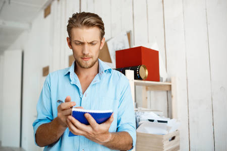 Portrait of young handsome confident pensive businessman writing in notebook. White modern office interior background.の写真素材