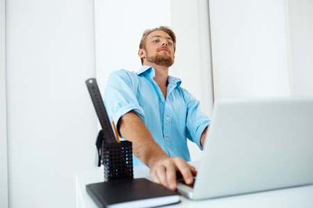 Picture of young handsome confident pensive businessman sitting at table working on laptop with cup...