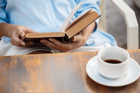 Hands of young girl sitting at the wooden table with book and coffee cup.