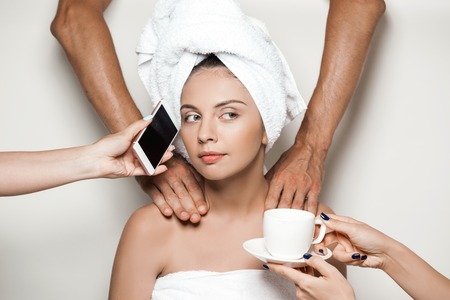 Hands doing massage, giving phone and cup of coffee to young beautiful naked girl in towel over light background.の写真素材