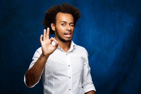 Young handsome african man in white shirt showing okay, winking over blue background. Copy space.の写真素材