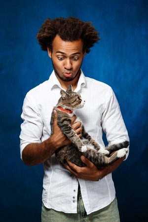Young handsome african man in white shirt holding cat over blue background. Copy space.