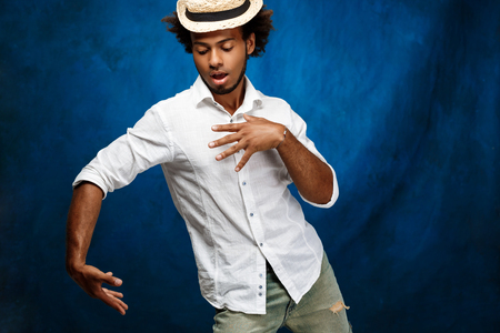 Young handsome african man in white shirt and hat dancing over blue background. Copy space.