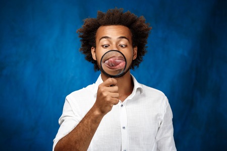 Young handsome african man in white shirt posing with magnifier over blue background. Copy space.