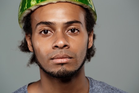 Young handsome frican man with watermelon on head fooling over grey background. Copy space.の写真素材