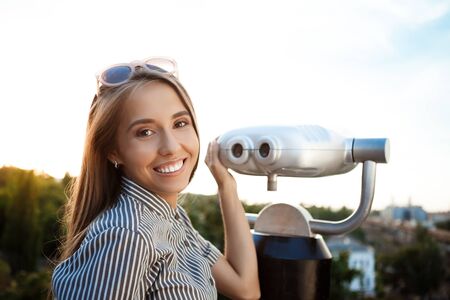 Young beautiful girl in sunglasses walking around city, posing near binoculars, smiling.
