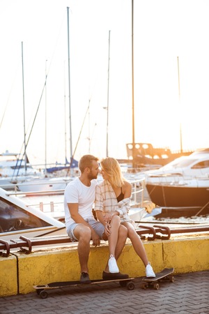 Young beautiful couple sitting at seaside, smiling, skateboarding. Outdoor backgroundの写真素材