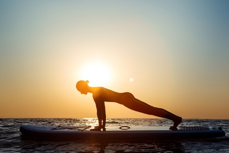 Silhouette of young beautiful girl practicing yoga on surfboard in sea at sunrise.