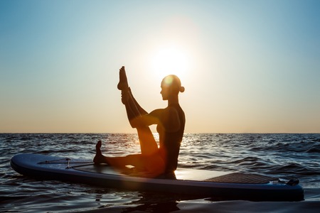 Silhouette of young beautiful girl practicing yoga on surfboard in sea at sunrise.の写真素材