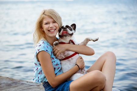Beautiful blonde girl smiling, sitting, playing with French bulldog near sea.の写真素材