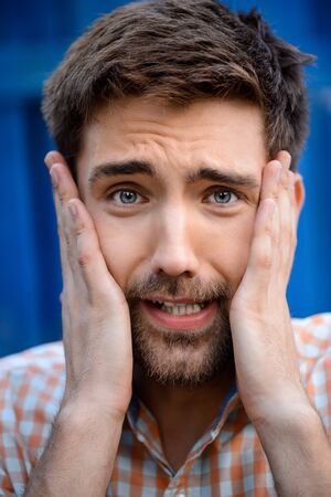 Close up portrait of handsome man upset holding face with hands, looking at camera over blue background.の写真素材