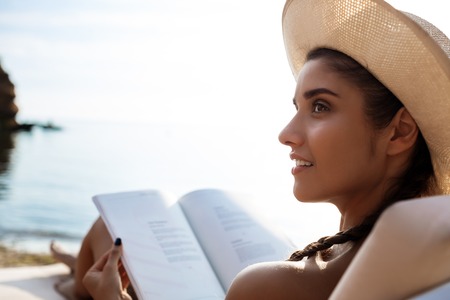 Beautiful brunette girl in hat smiling, reading book, lying at sea beach. Copy space.の写真素材