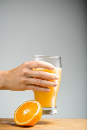 Girl s arm holding glass of juice over grey background. Half of orange lying on wooden desk. Copy space.の写真素材