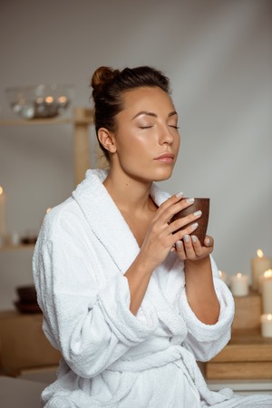 Young beautiful brunette girl holding tea cup relaxing in spa salon. Copy space.の写真素材