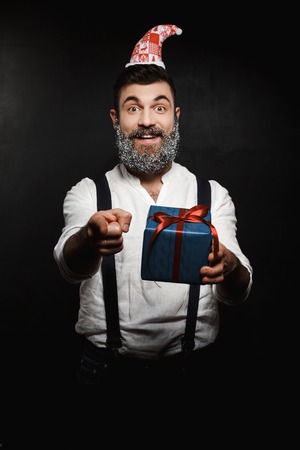 Young handsome man holding christmas gift box pointing finger at camera over black background. Copy space.の写真素材