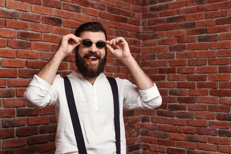 Young handsome man in sunglasses laughing holding cigar over brick background. Copy space.