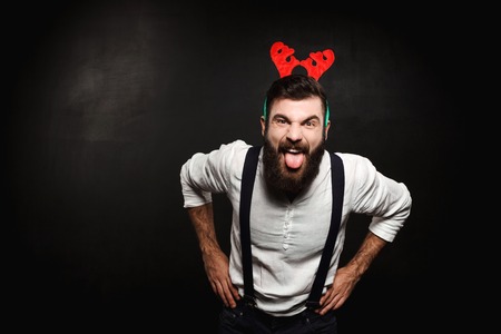 Young handsome man in fake deer horns showing tongue over black background. Copy space.