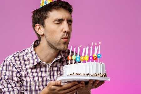 Young handsome man blowing out candles on birthday cake over purple background. Copy space.
