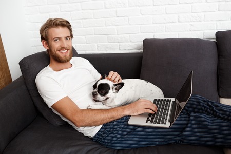 Young handsome man smiling looking at camera holding laptop stroking pug doglying on sofa.の写真素材