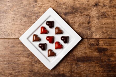 Chocolate candies in shape of heart on white plate over wooden background. From above. Copy space.