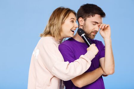 Young beautiful couple over blue background. Girl singing in microphone man didpleased. Copy space.の写真素材