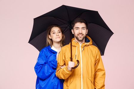 Young beautiful couple posing in rain coats holding umbrella over light pink background. Copy space.の写真素材