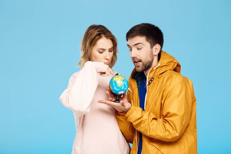 Young beautiful couple posing pointing at different places of small globe over blue background. Man wearing rain coat. Copy space.の写真素材