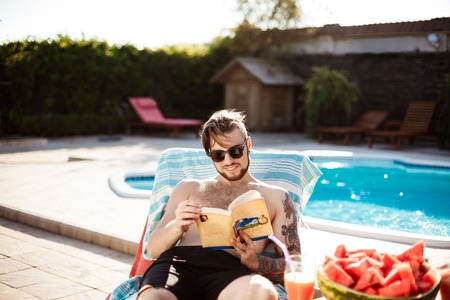 Handsome man reading book, lying on chaise near swimming pool.の写真素材