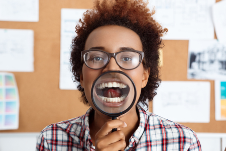 Young beautiful african worker holding magnifier infront of mouth. Office background.の写真素材