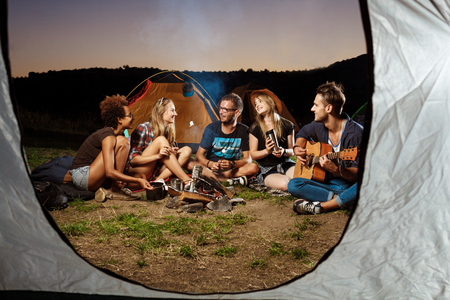Friends sitting near bonfire, smiling, playing guitar. Camping grill marshmallow.の写真素材