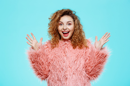 Close up portrait of cheerful surprised smiling beautiful brunette curly girl in pink fur coat over blue background. Copy space.の写真素材
