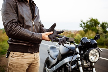 Young handsome man in leather jacket posing near his motorbike, wearing gloves at countryside road. Close up.の写真素材