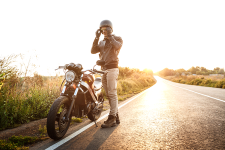 Young handsome man in leather jacket posing near his motorbike at countryside road.の写真素材