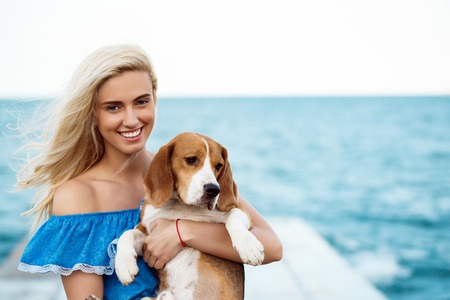 Young beautiful blonde girl in blue dress smiling, walking, playing with beagle dog at seaside.の写真素材